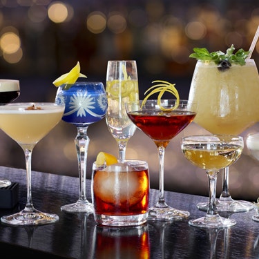 Cocktails i forskellige farver, typer og glas linet op på en bar.